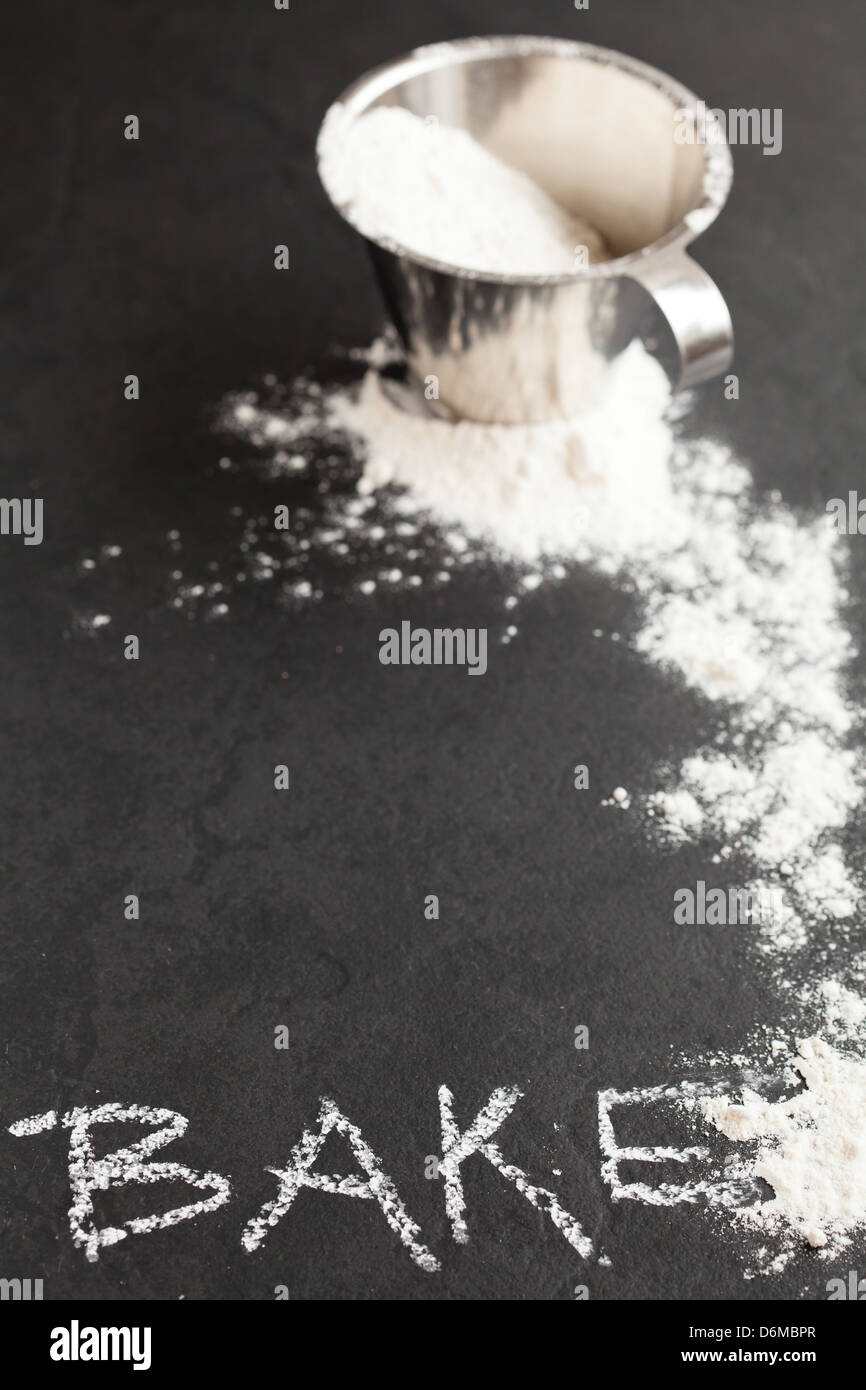 La harina en una taza medidora de metal y se extiende sobre un fondo oscuro con word bake escritos en tiza Foto de stock