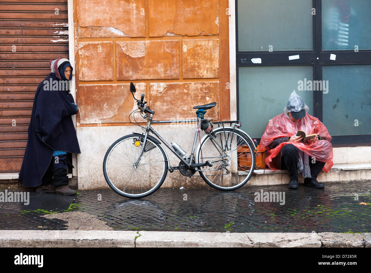 Mujer y un hombre sin hogar leyendo un libro en la lluvia están separados por una bicicleta Foto de stock