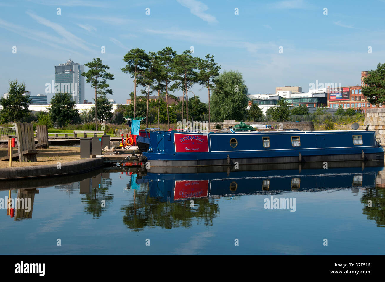 Barco narrowboat Canal y árboles reflejados en el puerto deportivo de Cotton Field Park, New Islington, Ancoats, Manchester, Inglaterra, Reino Unido. Torre de la ciudad en el fondo. Foto de stock