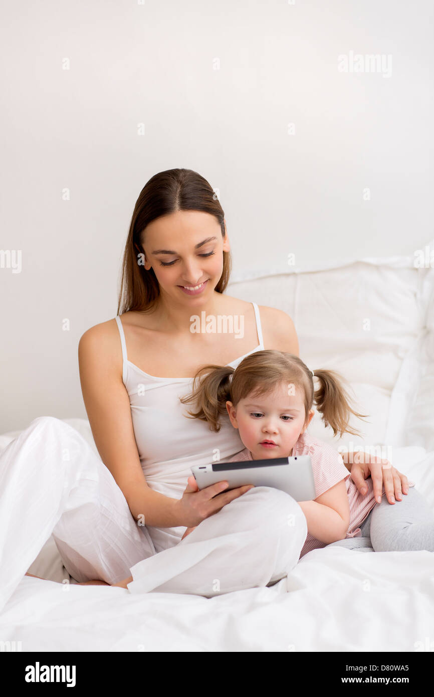 Niña jugando tableta digital con su mamá en el dormitorio blanco Foto de stock