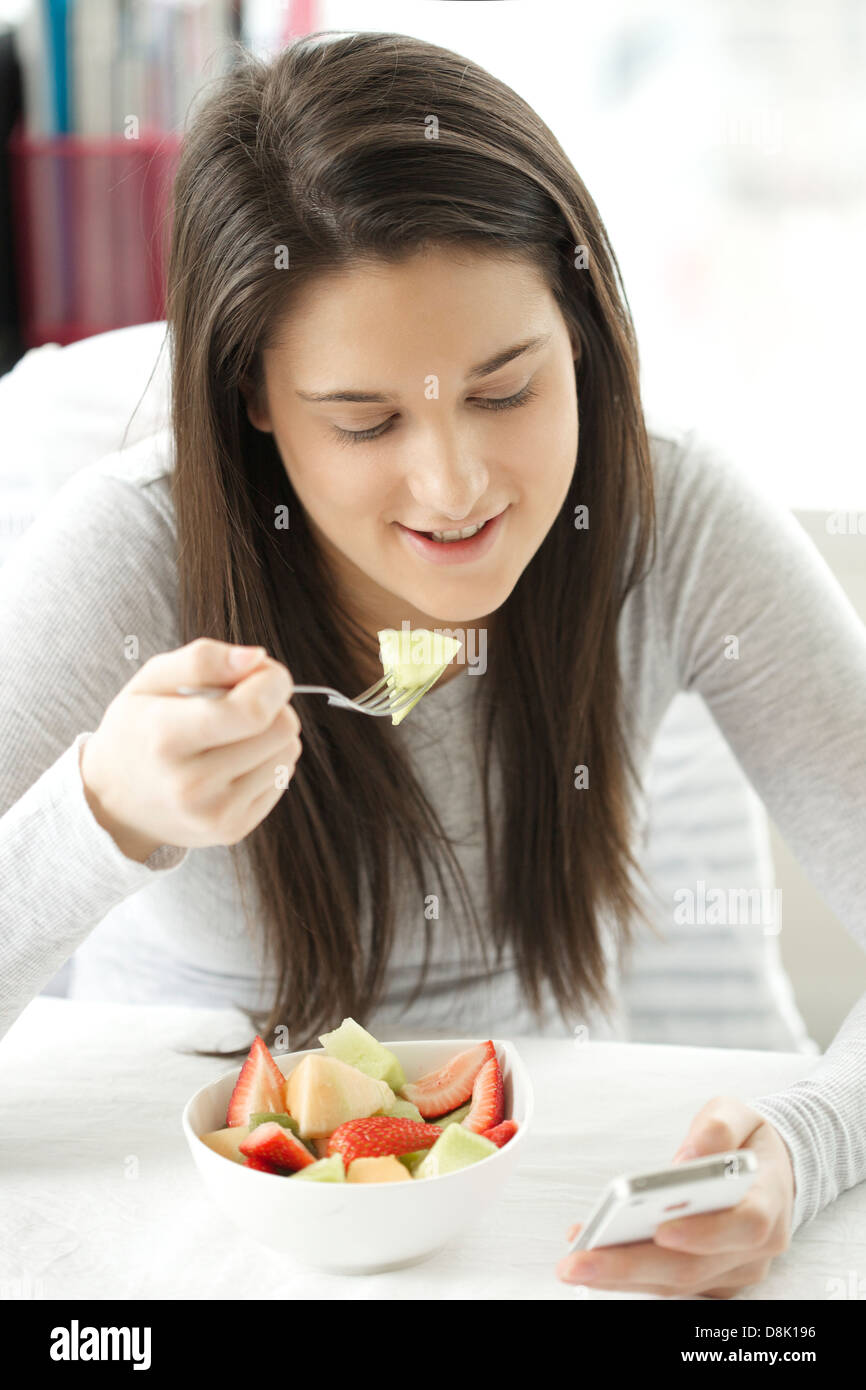 Retrato de una joven mujer comer ensalada de frutas mientras se navega con su iphone Foto de stock