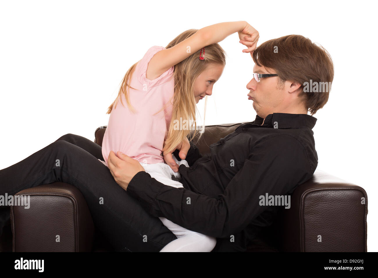 Padre joven jugar y hacer mueca a su hija y ella apuntando su dedo sobre su frente en un sillón de cuero marrón. Foto de stock