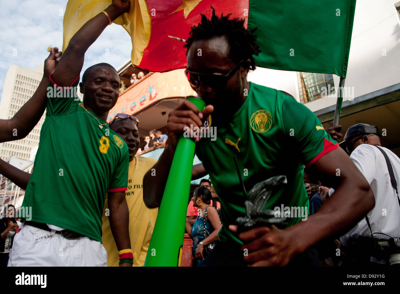 Un fanático del fútbol camerunés golpes vuvazula mientras amigo ondas bandera camerunesa por encima de él durante el sorteo final de la Copa Mundial de la FIFA Foto de stock