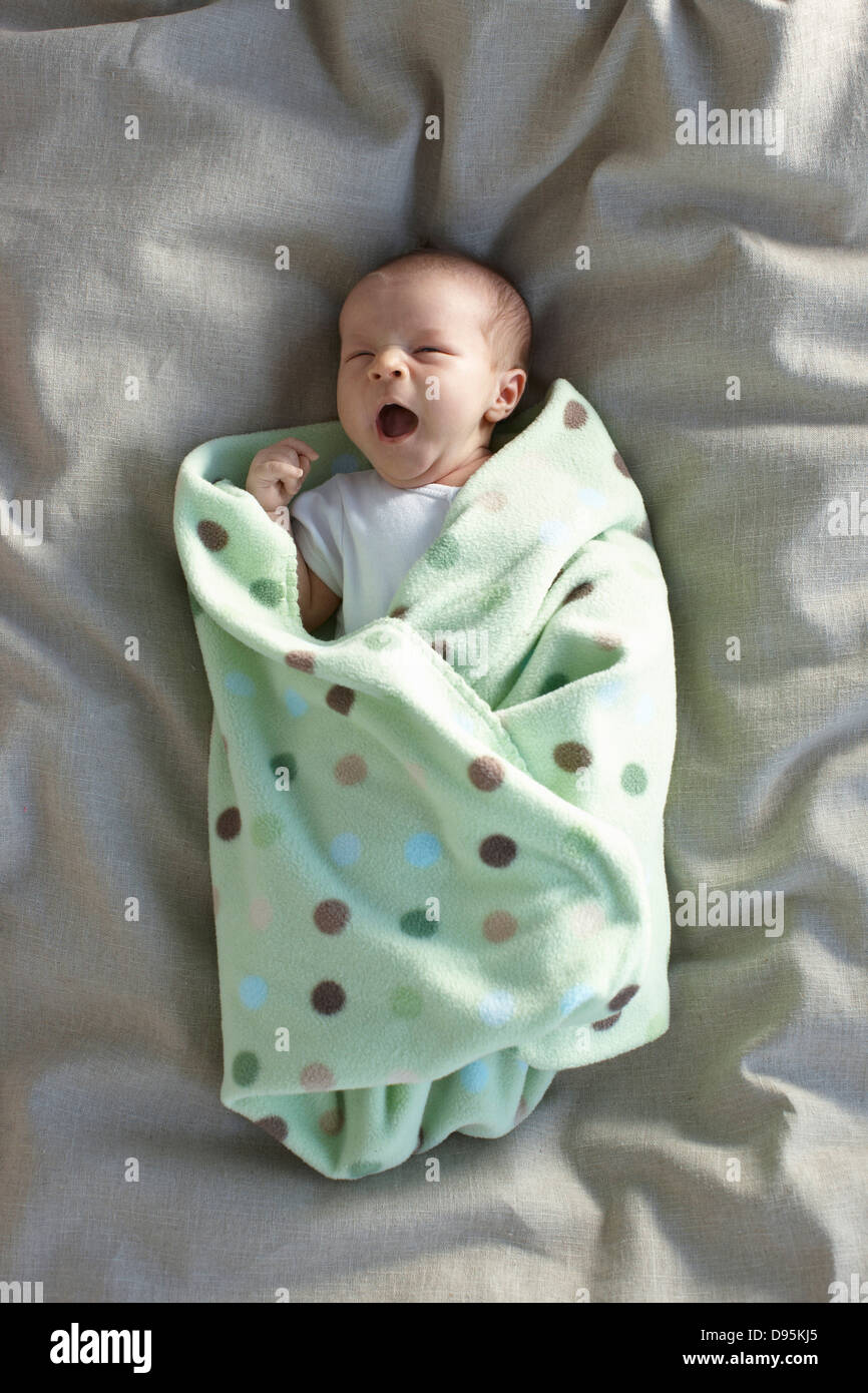 Bebé niña caucásica en una camiseta blanca sobre una cama swadled bostezo en una manta para bebés Foto de stock