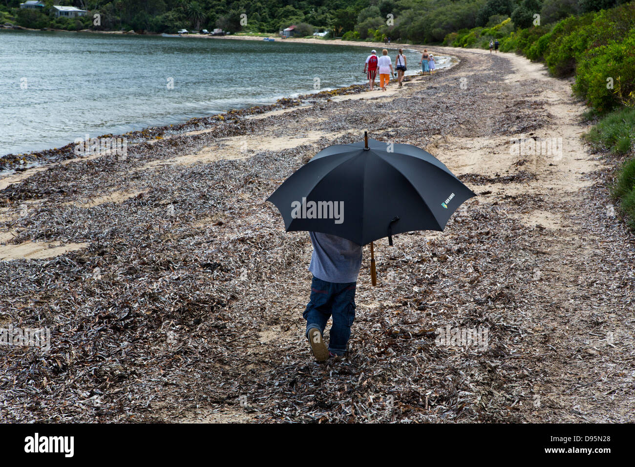 Pequeño niño en jeans de paseos a lo largo de la playa, bajo el paraguas de adultos Foto de stock