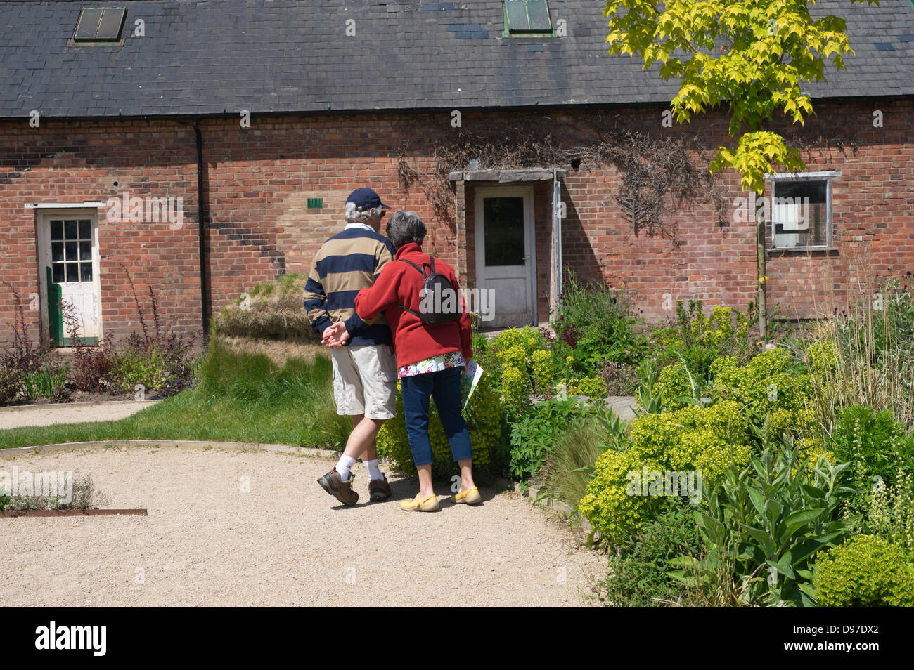 Buscando pareja senior y disfrutar de un jardín en el Reino Unido durante el verano, con un gran edificio de ladrillo rural en el fondo Foto de stock
