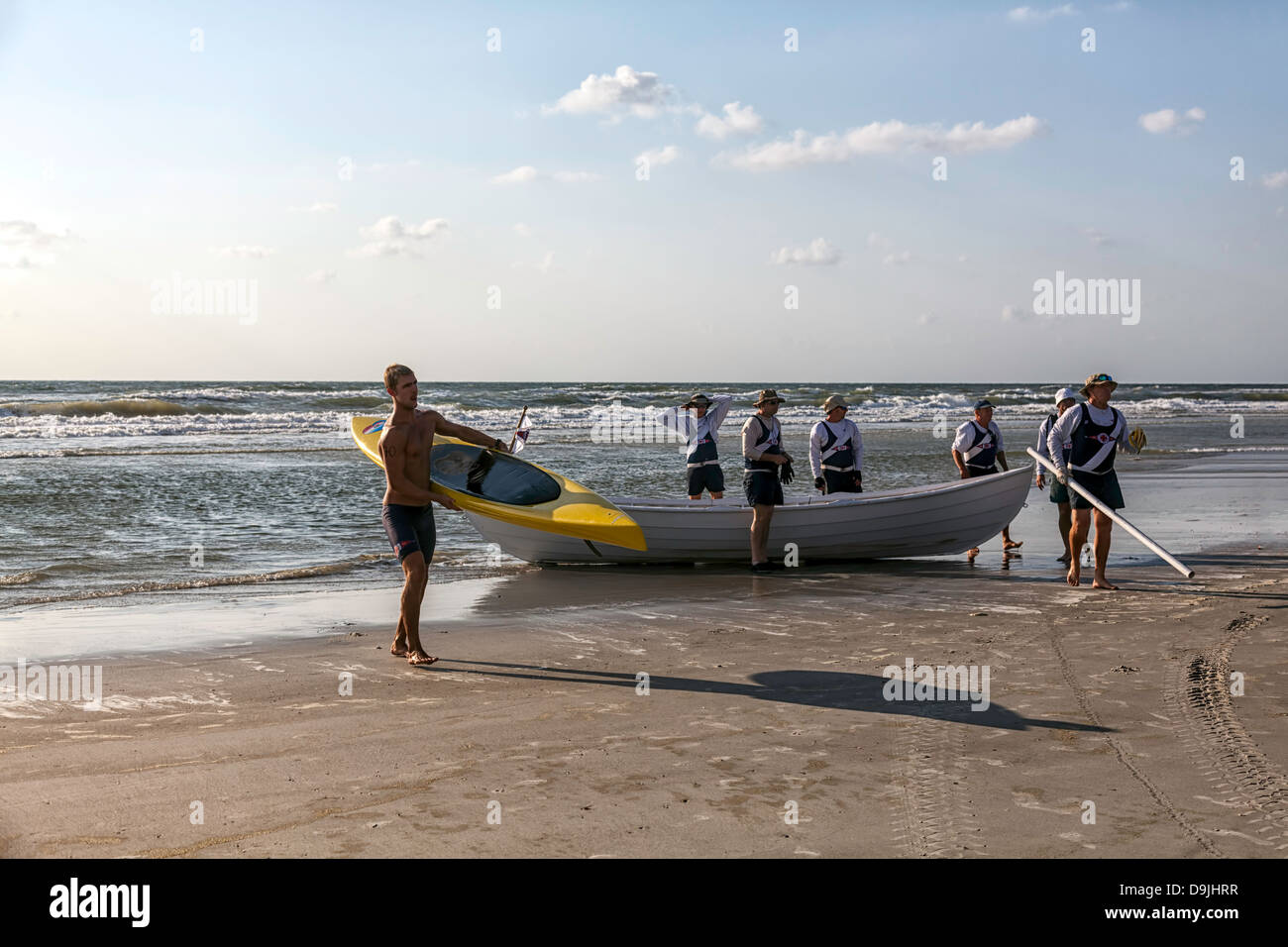 Hombre joven llevando su kayak de mar fuera del Océano Índico con bote de rescate y voluntarios en el fondo. Foto de stock