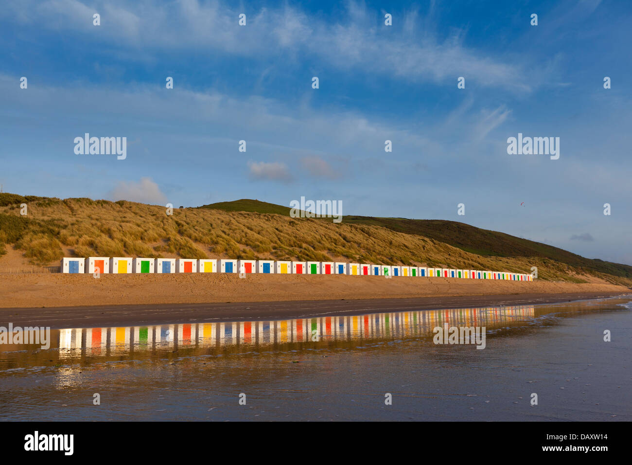 Una larga línea de cabañas de playa blanca, con puertas de colores, de pie delante de dunas con reflejos en la arena mojada. Foto de stock