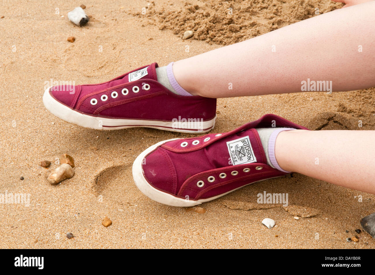 Un par de rojo oscuro laceless zapatillas sobre las piernas de una joven mujer en una playa de arena Foto de stock