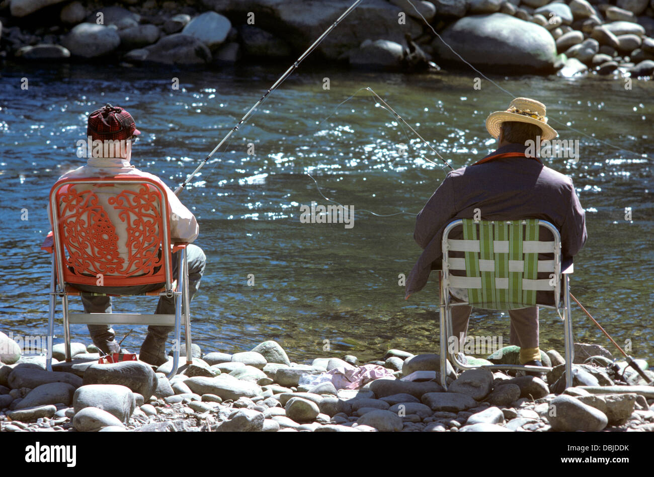 Dos pescadores jubilados relajarse en su silla de jardín a orillas del arroyo de la montaña rocosa en la década de los 70 Foto de stock