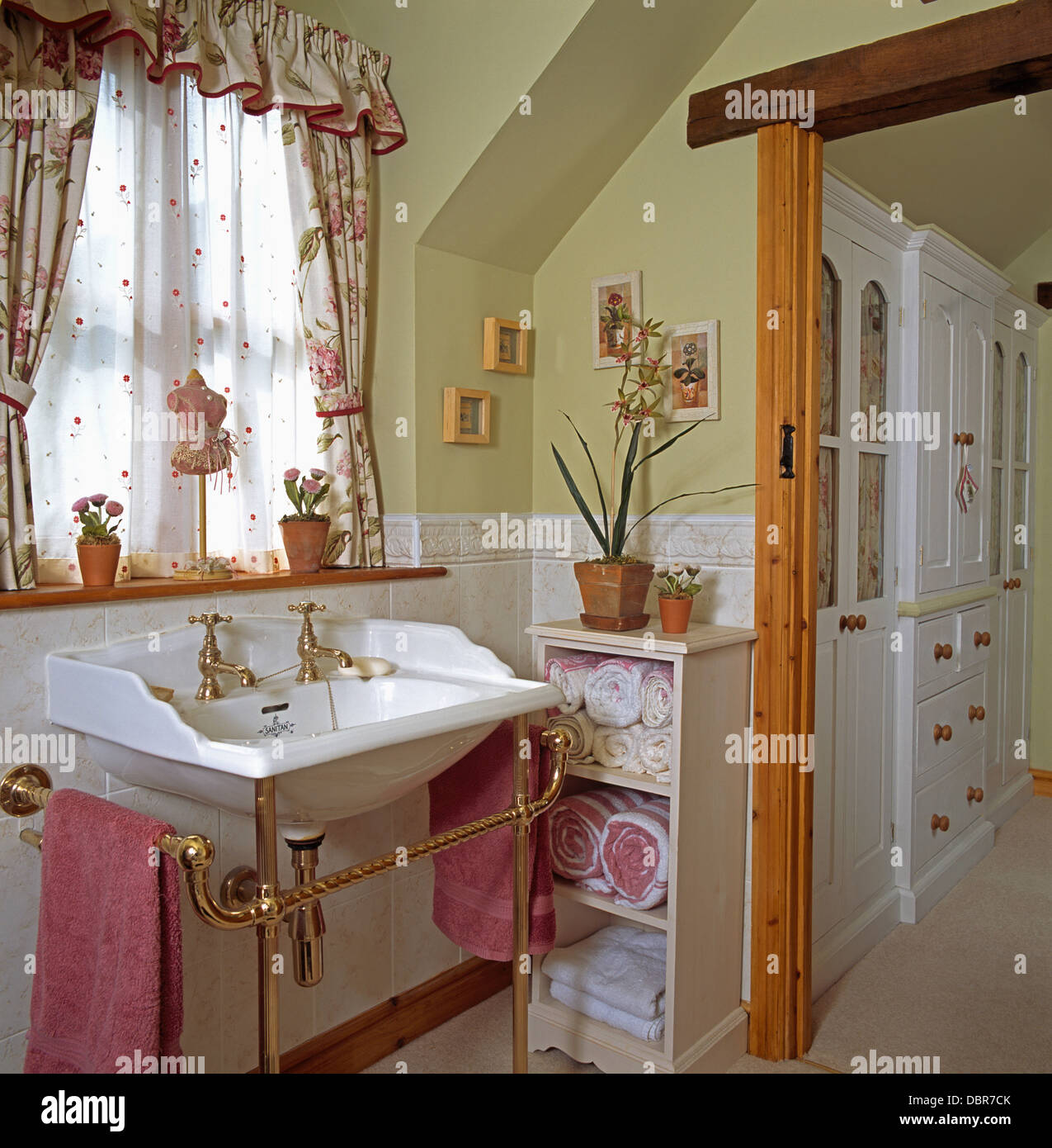 De velo blanco y cortinas con motivos florales en la ventana anterior en la cuenca de cuarto de baño privado con toallas enrolladas almacenados en estantería Foto de stock