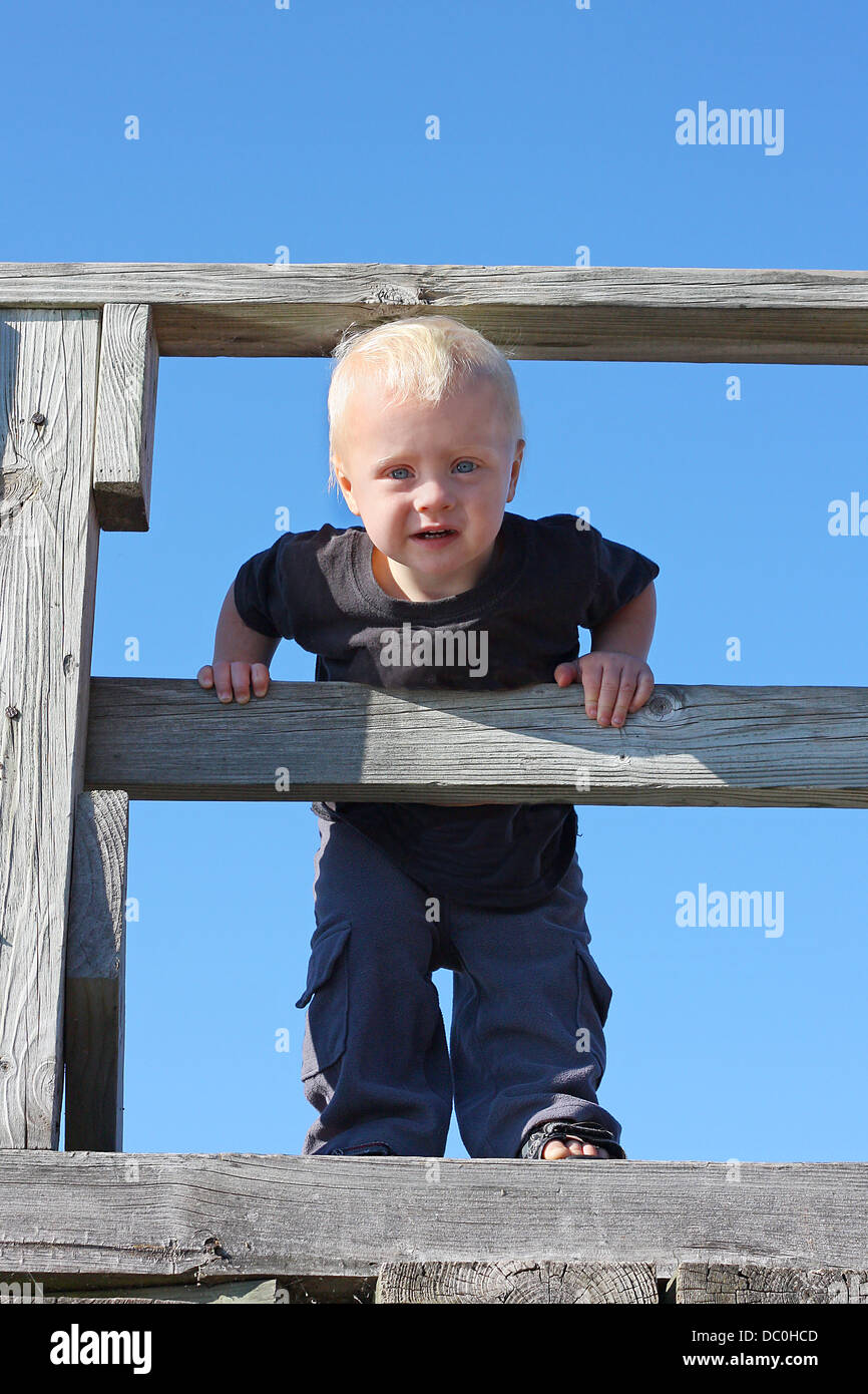 Un lindo Baby Boy es la escalada en la baranda de madera de un muelle en Playa del Lago Superior en un día soleado de verano. Foto de stock