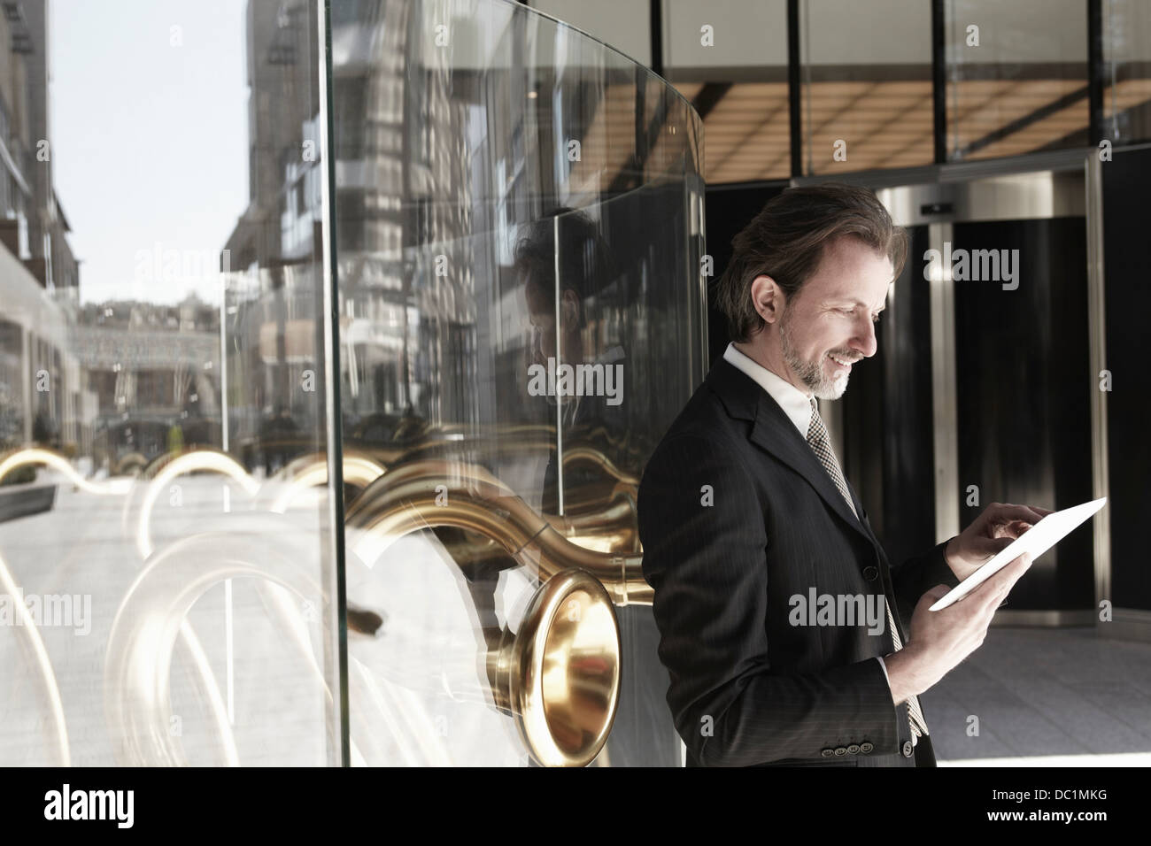 Adulto medio empresario apoyado contra la ventana de guardar y usar tableta digital Foto de stock