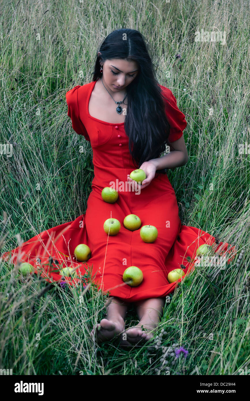 Una mujer en un vestido rojo sentado en un campo, con manzanas verdes en su vestido Foto de stock