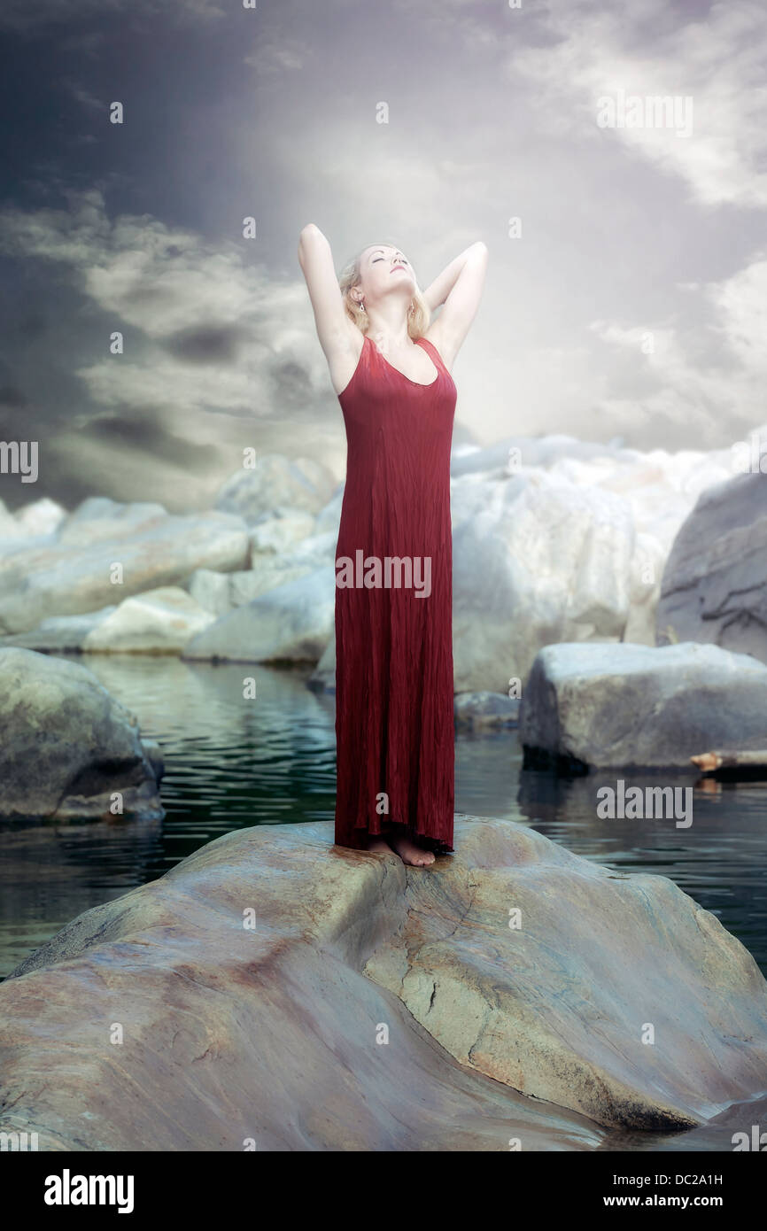 Una mujer en un vestido rojo está de pie sobre una roca en el agua y disfrutar del sol Foto de stock