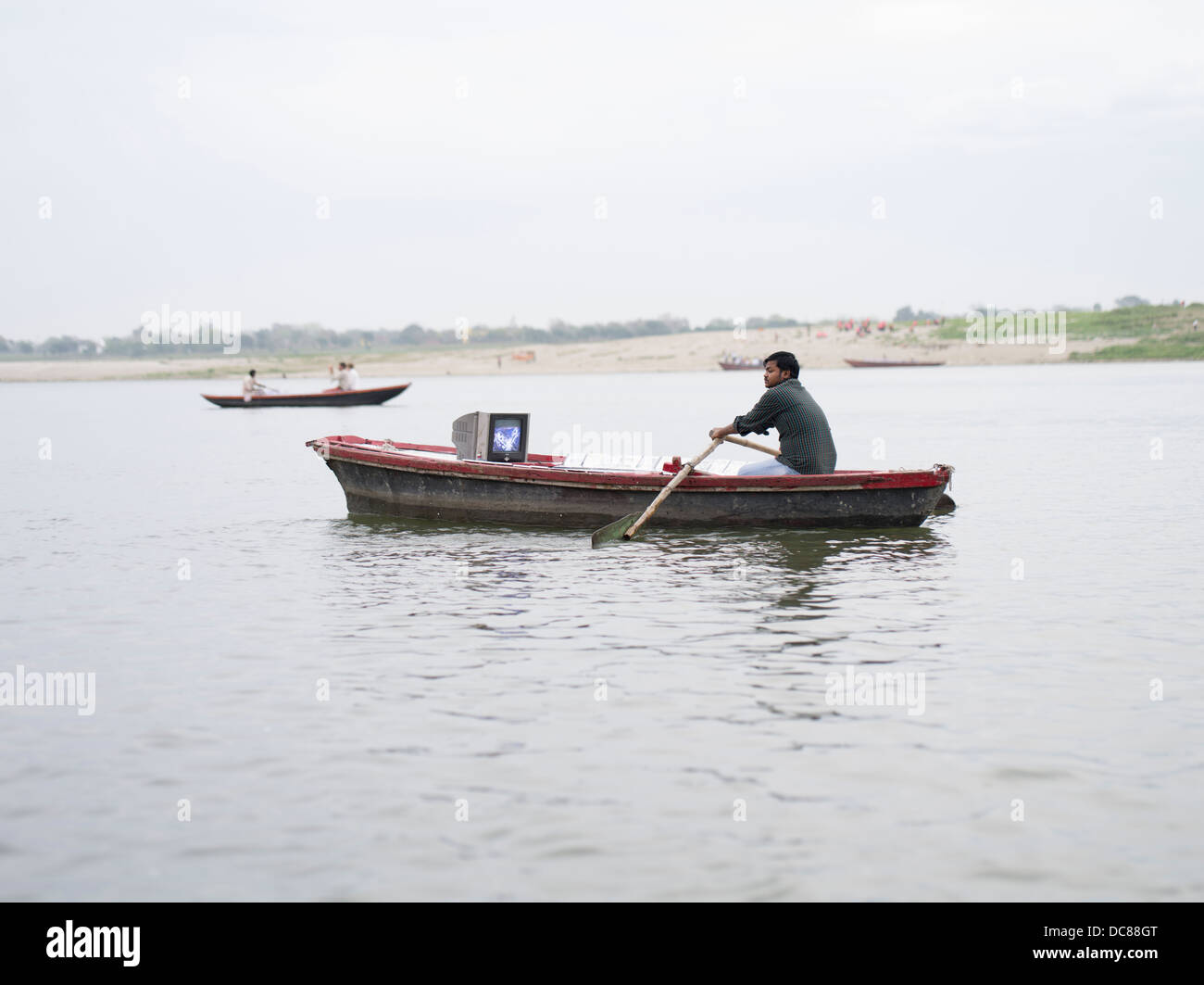 Hombre con TV dentro de su bote de remos. Venta de DVD sobre el río Ganges - Varanasi, India Foto de stock