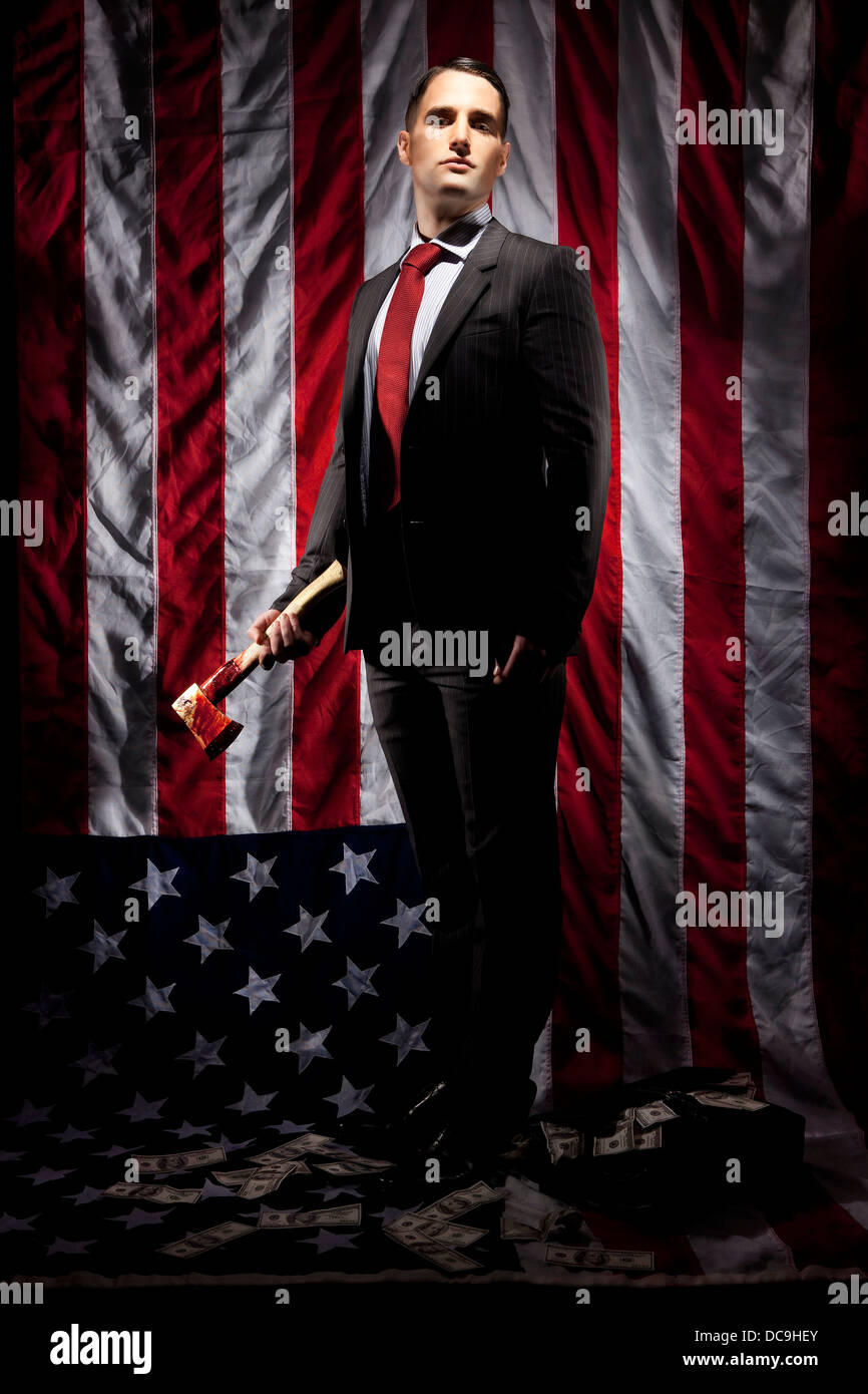 Un banquero sosteniendo un hacha sangrienta con billetes de cien dólares alrededor de sus pies, de pie sobre una bandera americana, planteados por el modelo. Foto de stock