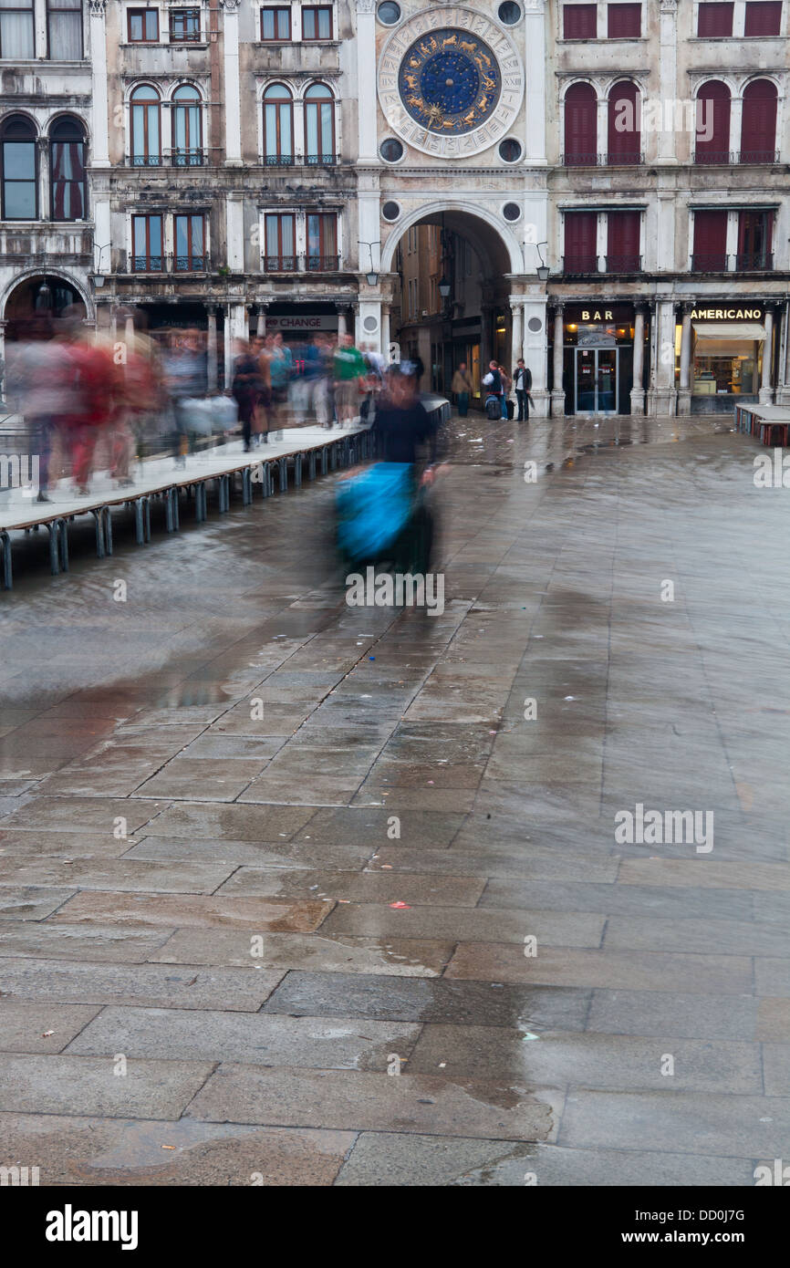 Aqua Alta. inundaciones en Piazza San Marco: el desenfoque de movimiento de peatones caminando sobre duckboards, hombre wheeling carretilla, cubierto de azul Foto de stock