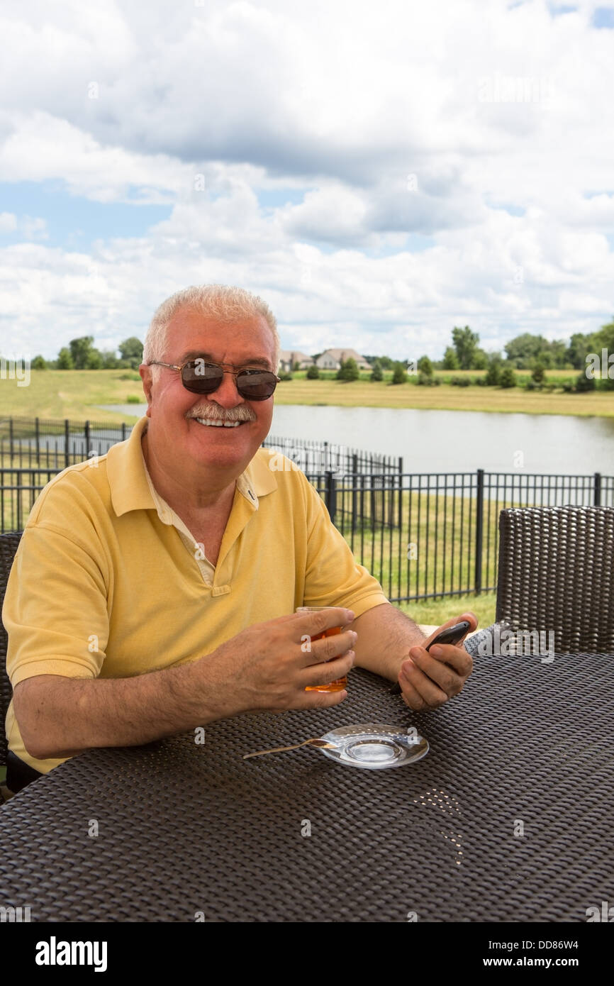 Hombre Senior posando con su teléfono inteligente mientras bebe su té turco en el patio alrededor del estanque. Foto de stock