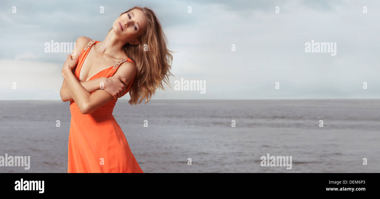 Joven y bella mujer sonriente en naranja vestido de verano de pie en una orilla del mar Foto de stock