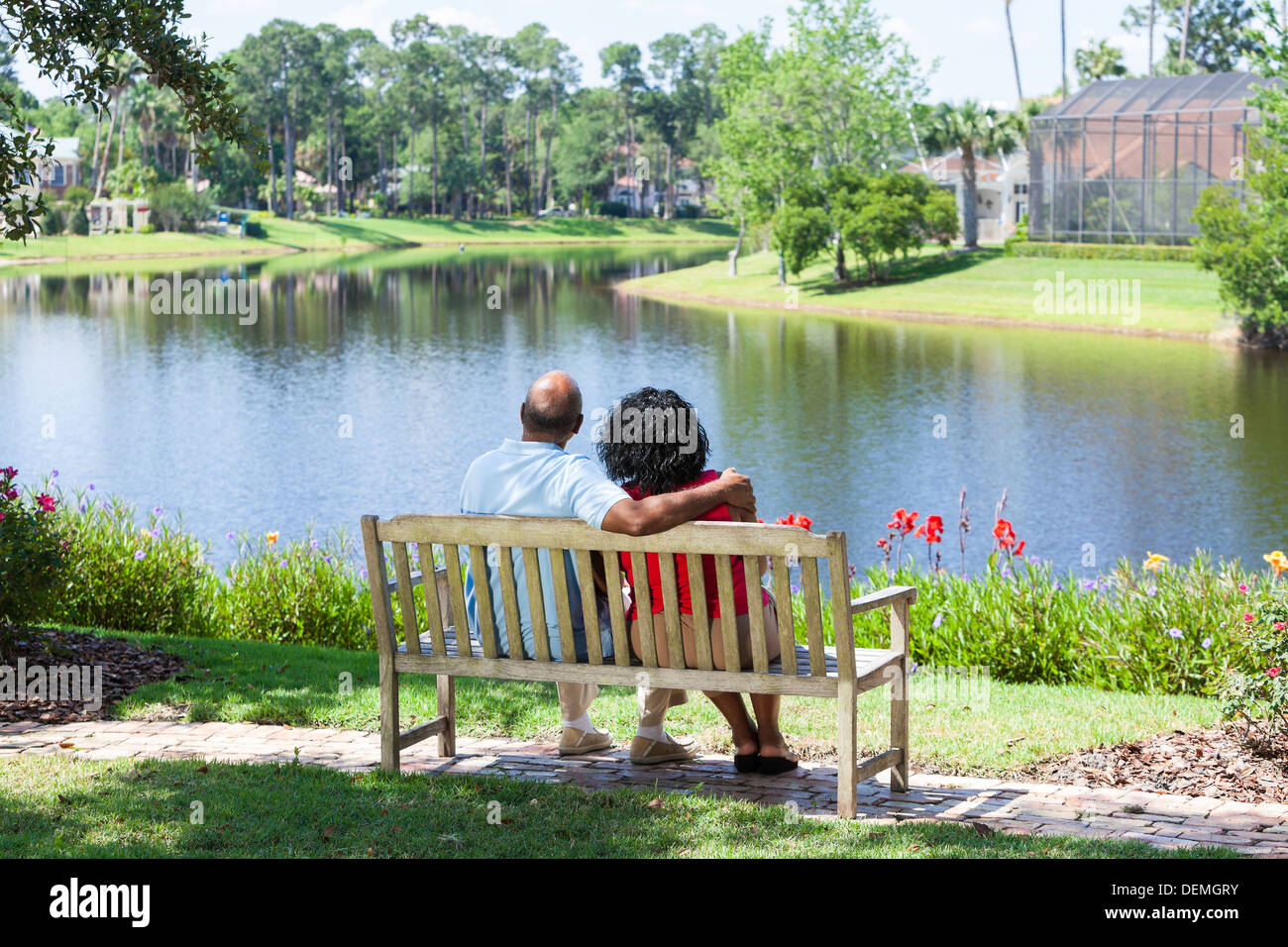 Vista trasera de un alegre romántica senior pareja Afroamericano sentado en una banca del parque abrazando mirando un lago Foto de stock