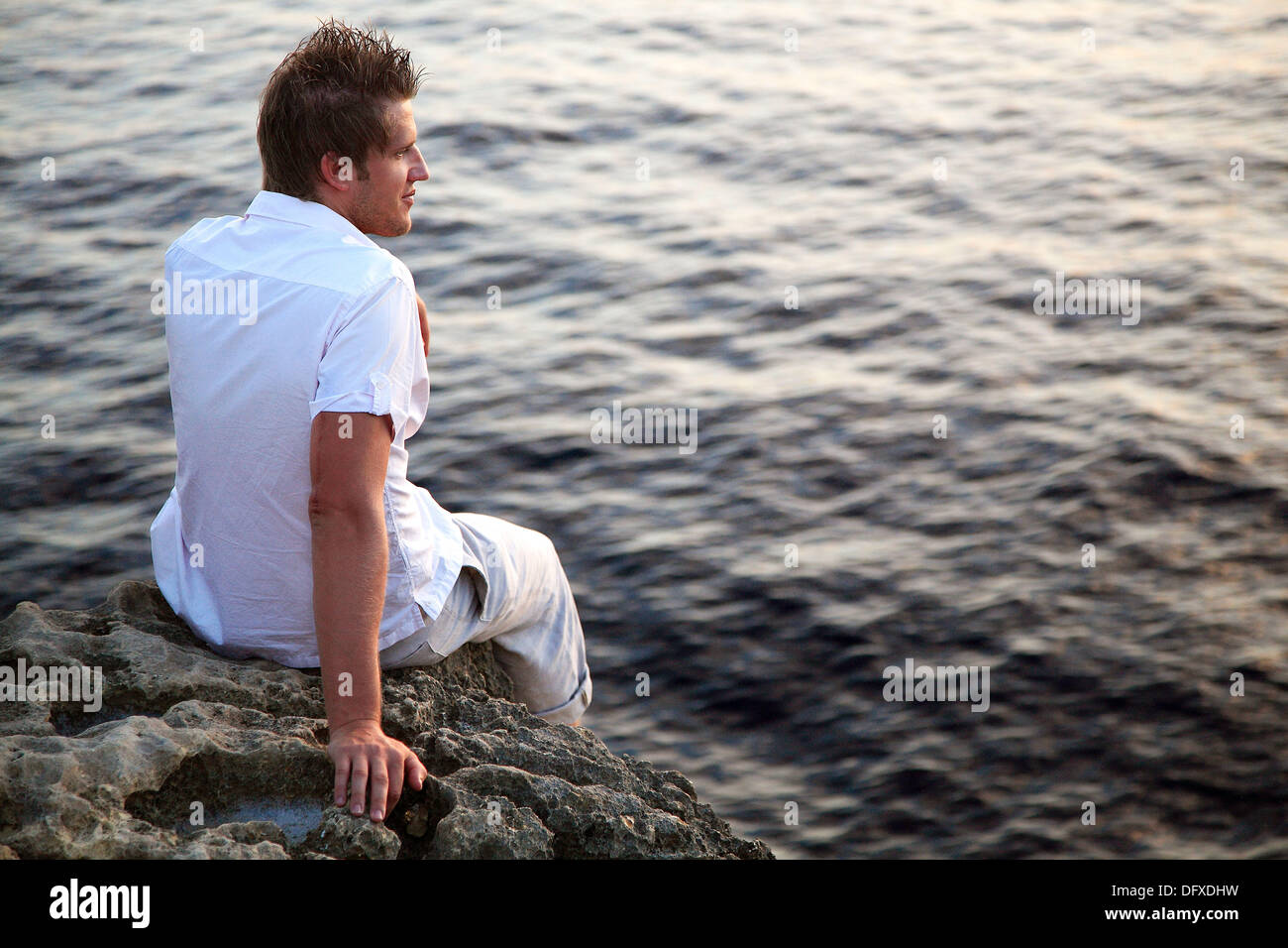 Curtidos hombres caucásicos con el pelo castaño oscuro, de 25 años, vestido con camisa blanca mira a través de una puesta de sol sobre el mar en Dwerja, en la isla de Foto de stock