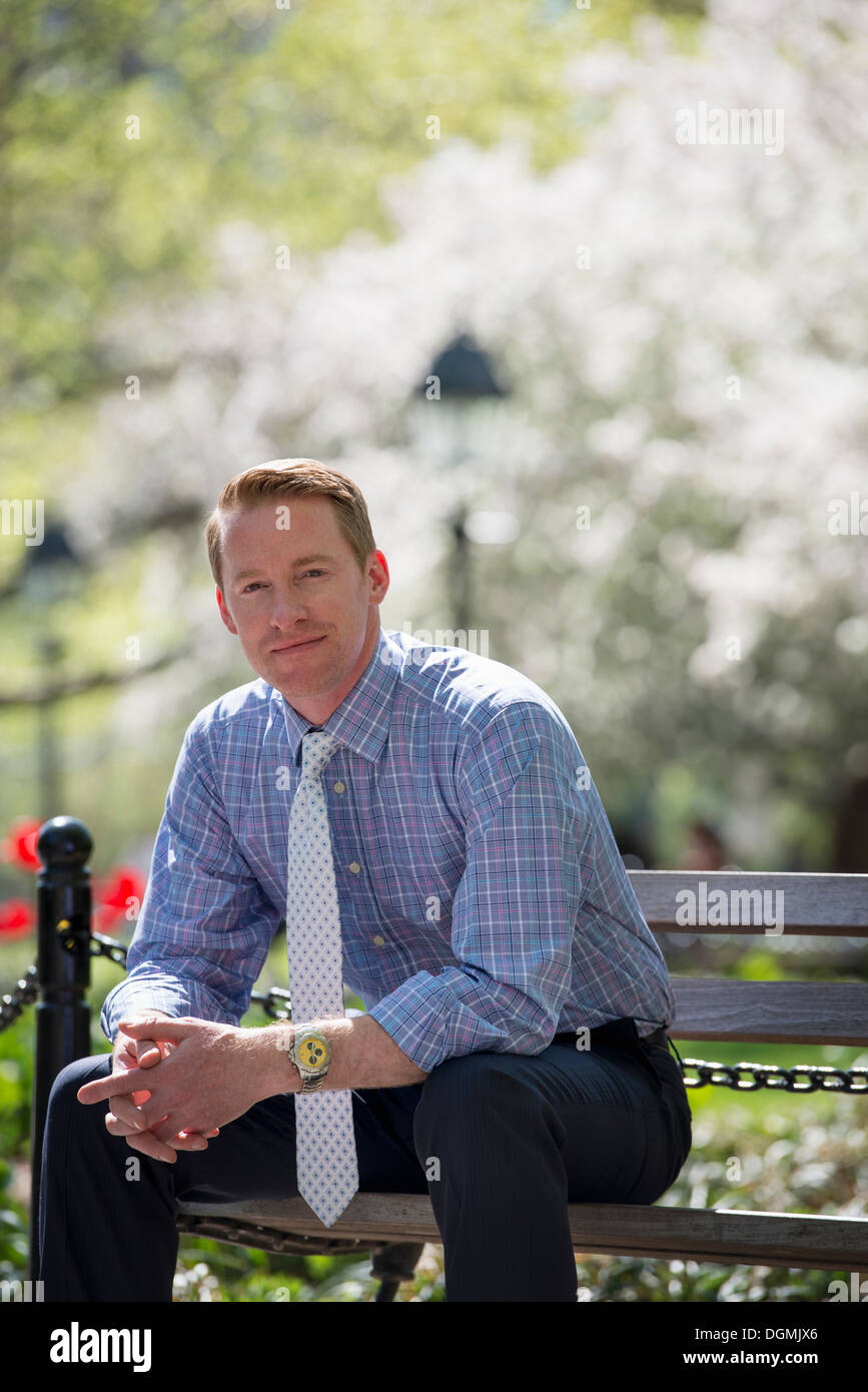 Un empresario en una camisa con corbata blanca, sentado en un banco del parque bajo la sombra de un árbol en flor. Foto de stock