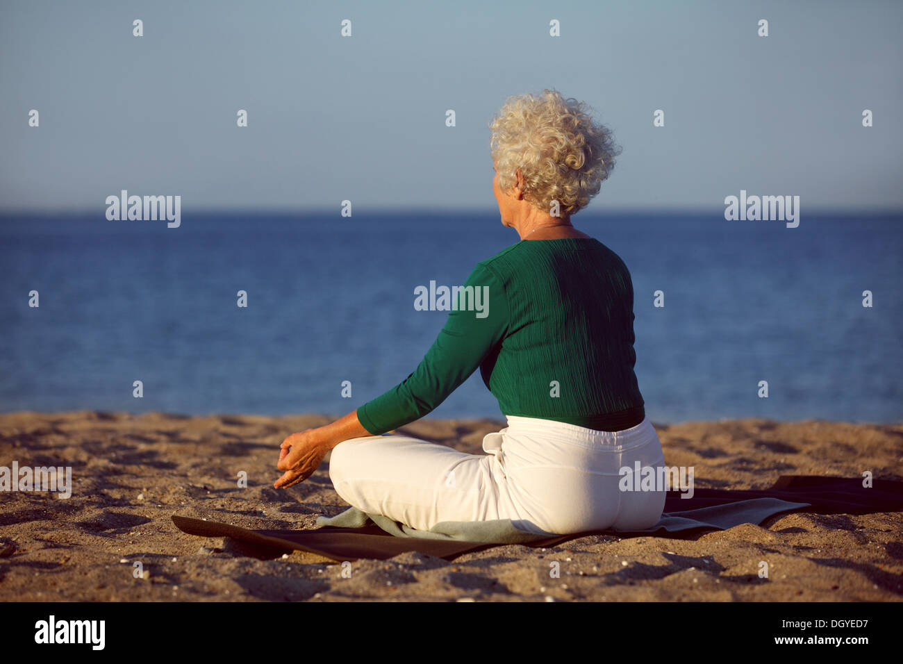 Vista lateral de la mujer de más edad en la meditación en la playa. Anciana sentado en la playa en lotus plantean haciendo ejercicio de relajación Foto de stock
