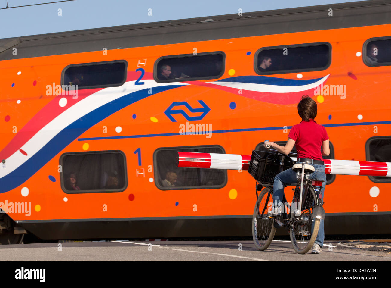 Países Bajos, Vogelenzang, tren paso a nivel. Mujer en bicicleta esperando la barrera. Naranja Royal Kings pasando por tren. Foto de stock