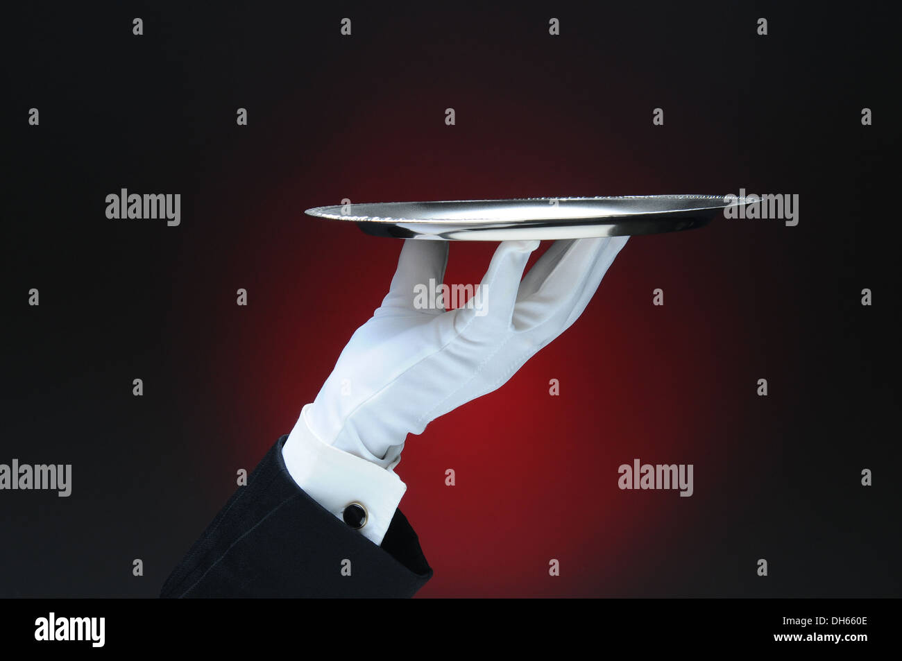 Primer plano de la mano de un camarero sosteniendo una bandeja de servicio de plata en la punta de sus dedos a través de una luz de fondo rojo oscuro. Foto de stock