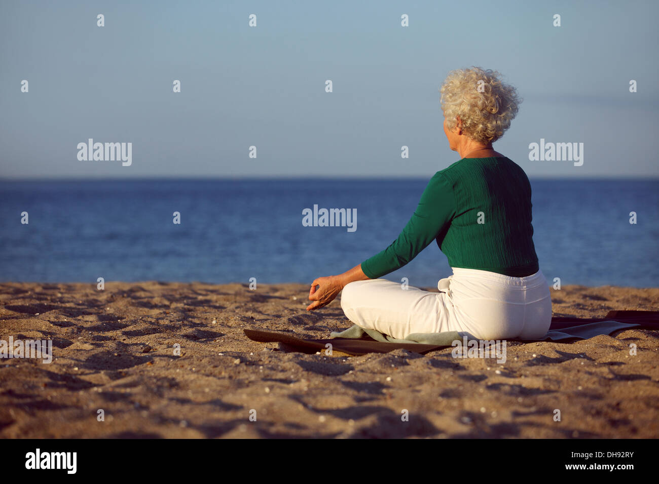 Vista lateral de la anciana en la meditación en la playa. Senior señora sentada en la playa en lotus plantean haciendo ejercicio de relajación. Foto de stock