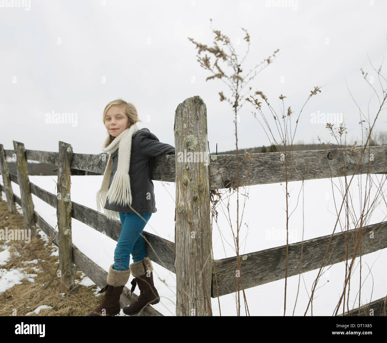 Una granja en la nieve, una niña con un sombrero y una bufanda y botas, apoyándose en una valla Foto de stock