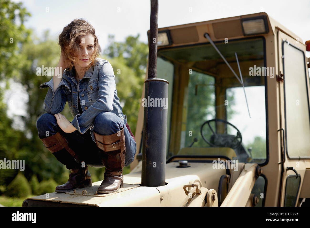 Una mujer joven en jeans chaqueta y botas sobre el capó del tractor Foto de stock