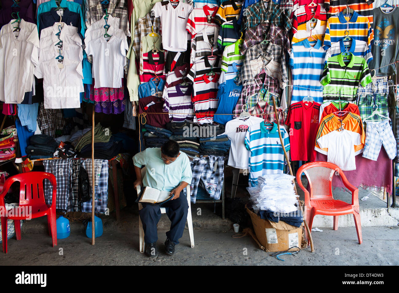 Un hombre lee un periódico sentado esperando para vender ropa en un puesto en el departamento de La Libertad, una ciudad portuaria en la costa de El Salvador Fotografía de -