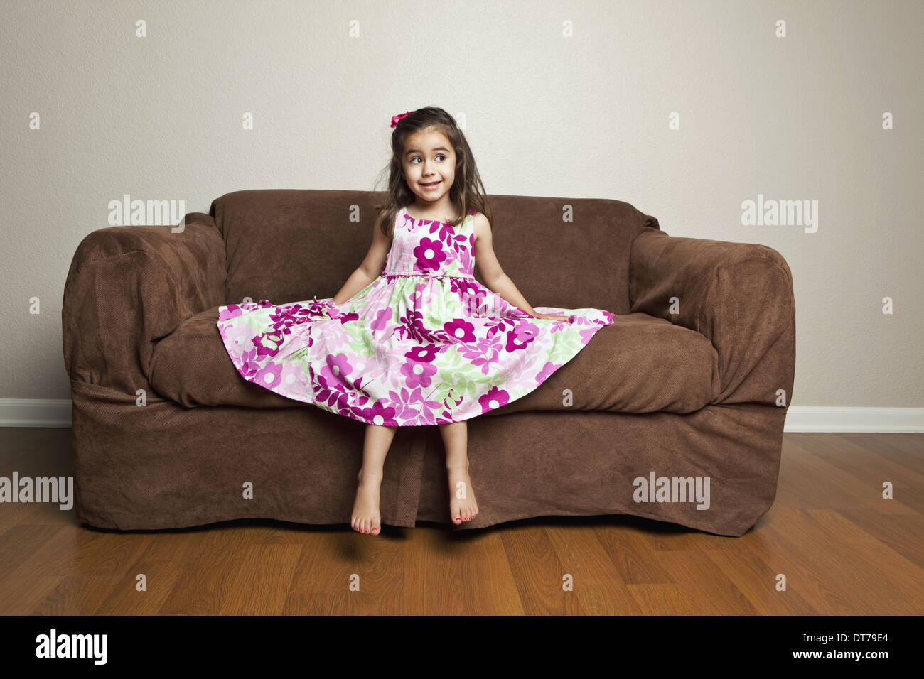 Una niña de 3 años de edad con el cabello marrón largo en un vestido de  algodón con flores rosas con la falda extendida, sentado en un sofá de  color marrón Fotografía