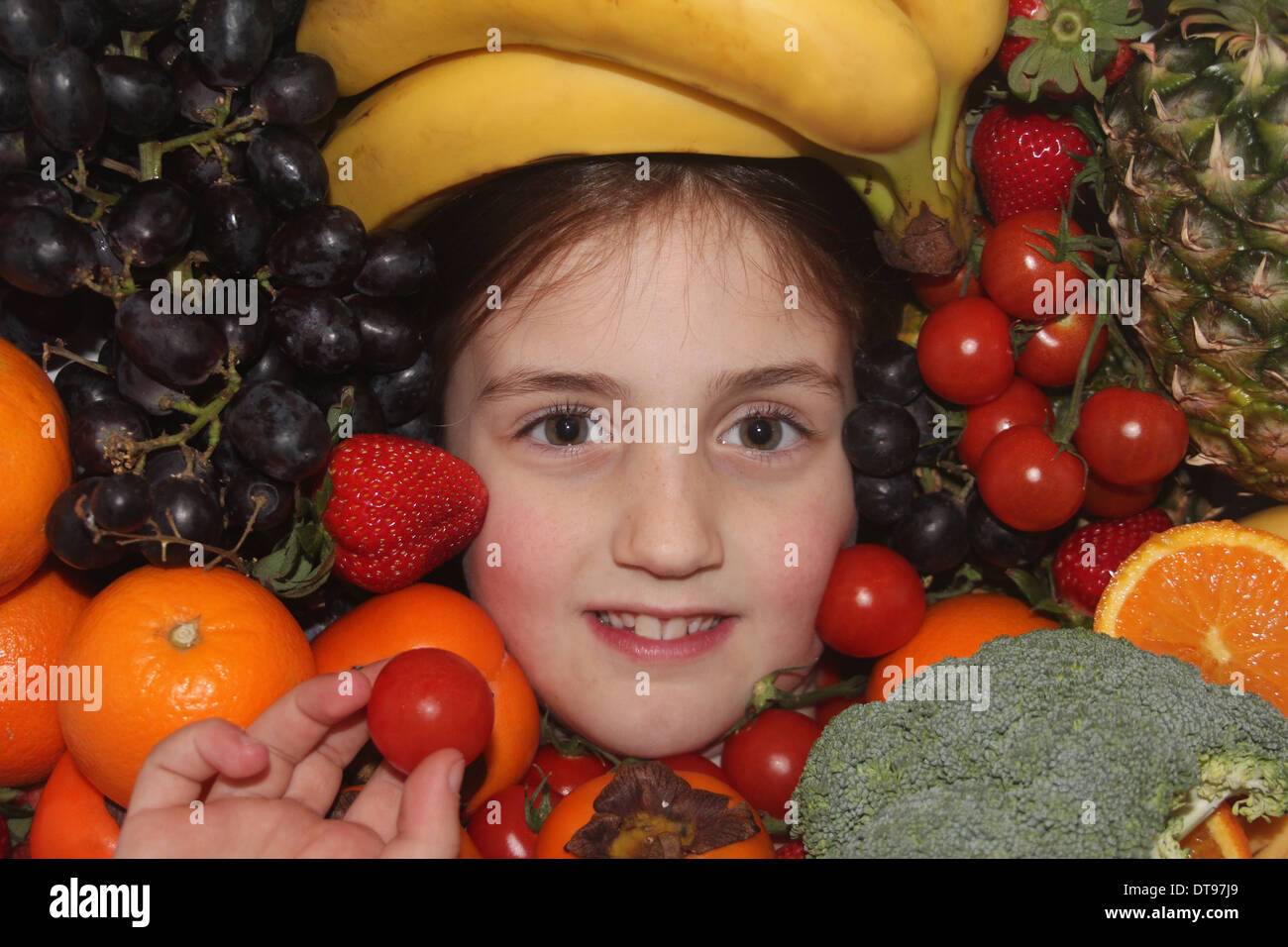 Los jóvenes caucásicos cara de la niña rodeada de frutales y hortalizas sosteniendo un tomate cherry, cinco al día, Inglaterra, Reino Unido. Foto de stock