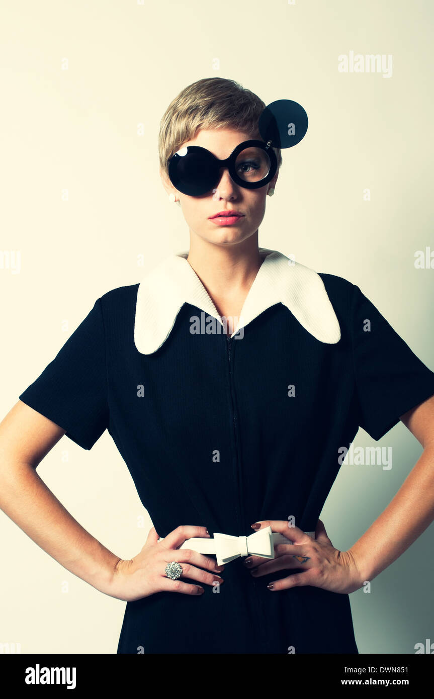 Una mujer retro posando en un mod 60s vestido, posando con gafas de sol  redonda plegable, con las manos en la cadera, un retro vintage 60s fashion  concept Fotografía de stock -