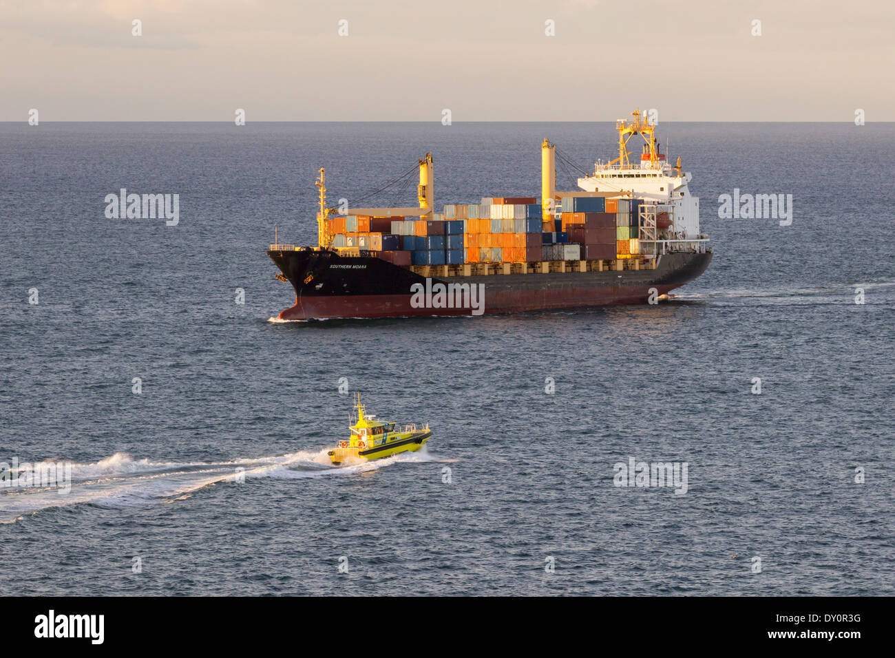 La carga de un buque portacontenedores con barco piloto amarillo acercándose al atardecer Foto de stock