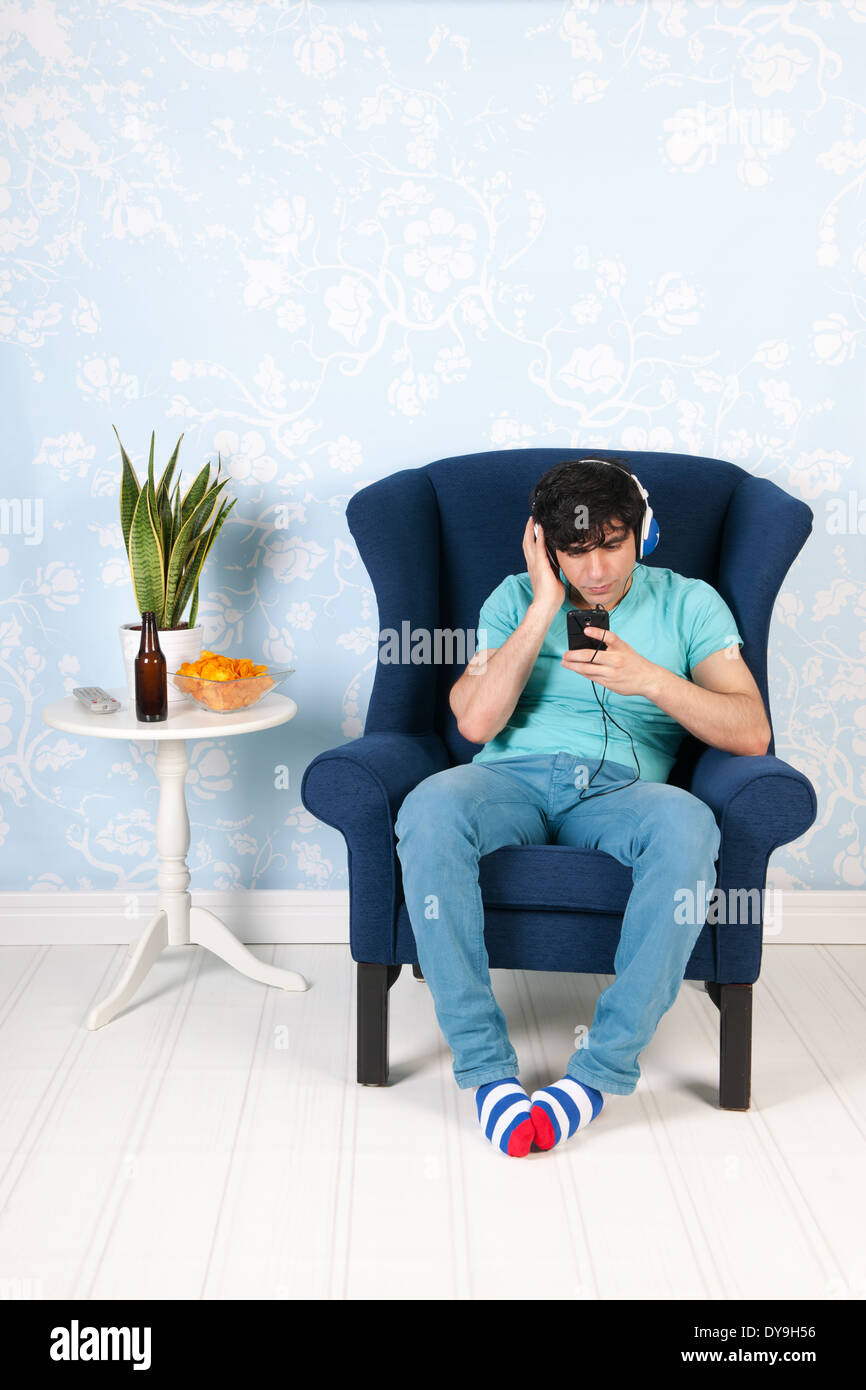 Hombre joven sentado en el living escuchando música con el teléfono inteligente. Foto de stock