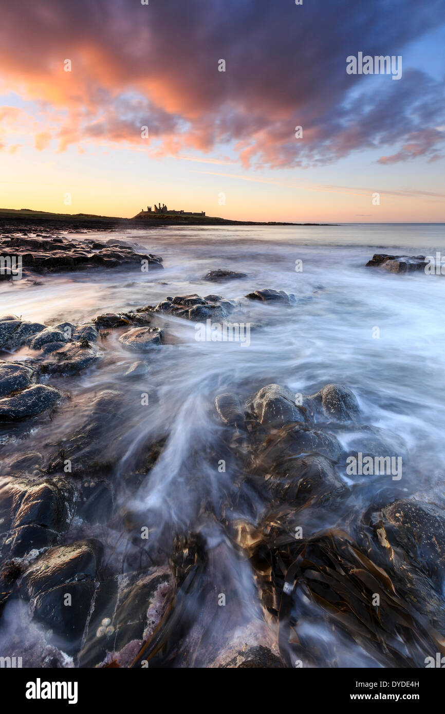 Atardecer en el castillo de Dunstanburgh con olas rompiendo sobre las rocas en primer plano. Foto de stock