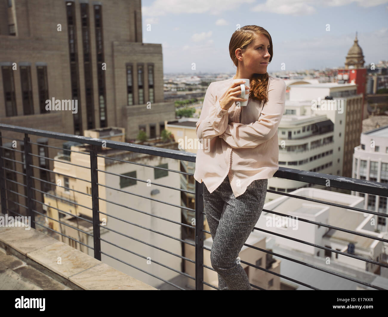 Hermosa señorita de pie por una verja en la terraza sosteniendo una taza de café mirando a otro lado. Mujeres caucásicas bebiendo café. Foto de stock