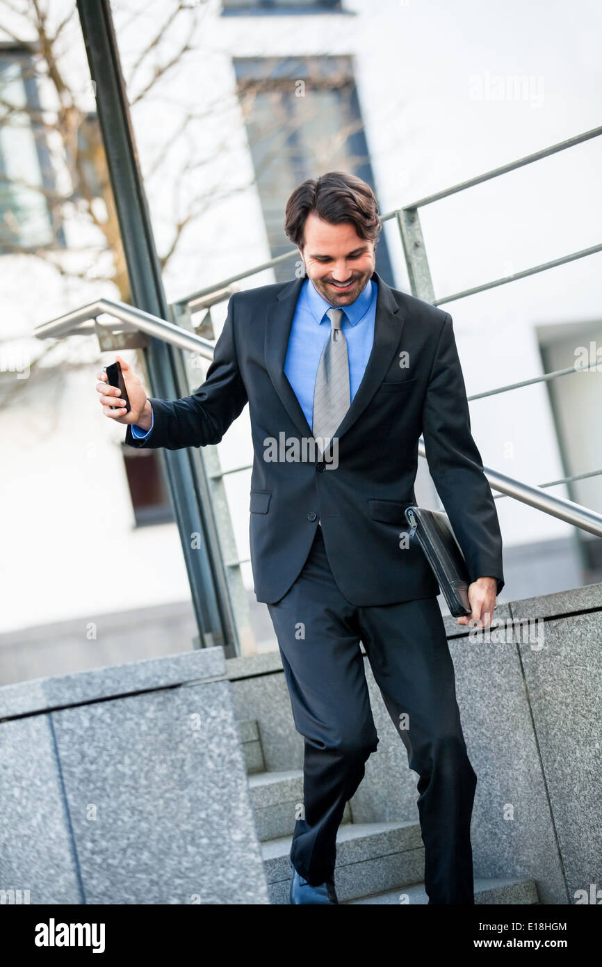 Elegante y sonriente joven empresario caminando por un tramo de escaleras en un entorno urbano al aire libre la celebración de un maletín o un folio en hi Foto de stock