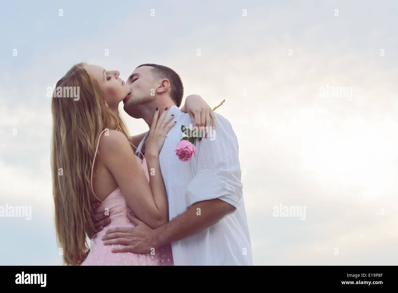 Una joven pareja romántica playa del retrato al atardecer besos. La mujer lleva un vestido vintage 50s, sosteniendo una rosa rosa. Foto de stock
