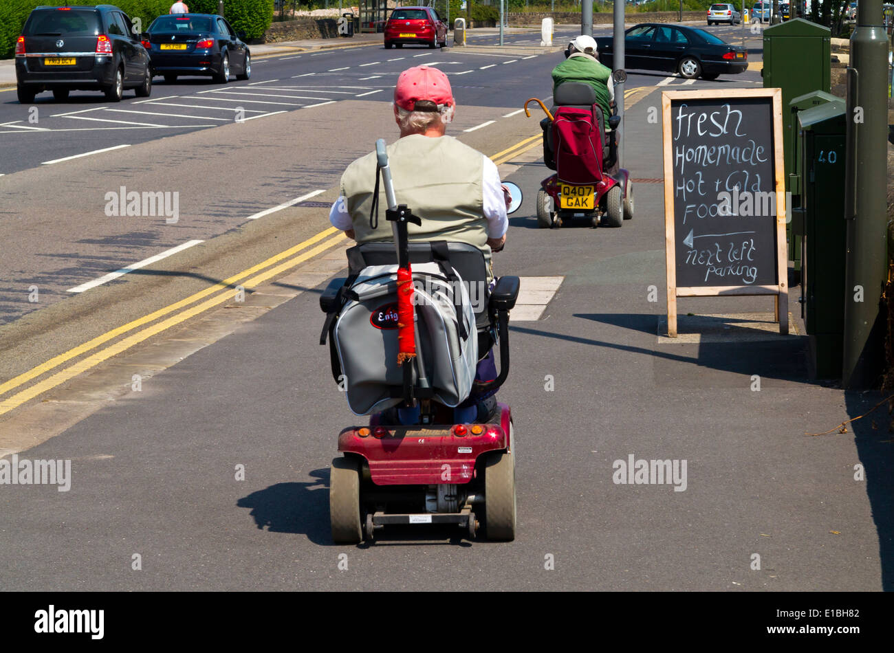 Dos personas de la tercera edad de equitación scooters de movilidad a lo largo de un pavimento de Chesterfield, Derbyshire, Inglaterra Foto de stock