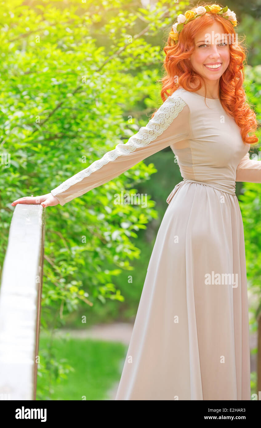 Hermosos vestidos de novia feliz largo vestido beige y corona de flores en  el pelo rojo, de pie en el pequeño puente en verano fresco park Fotografía  de stock - Alamy