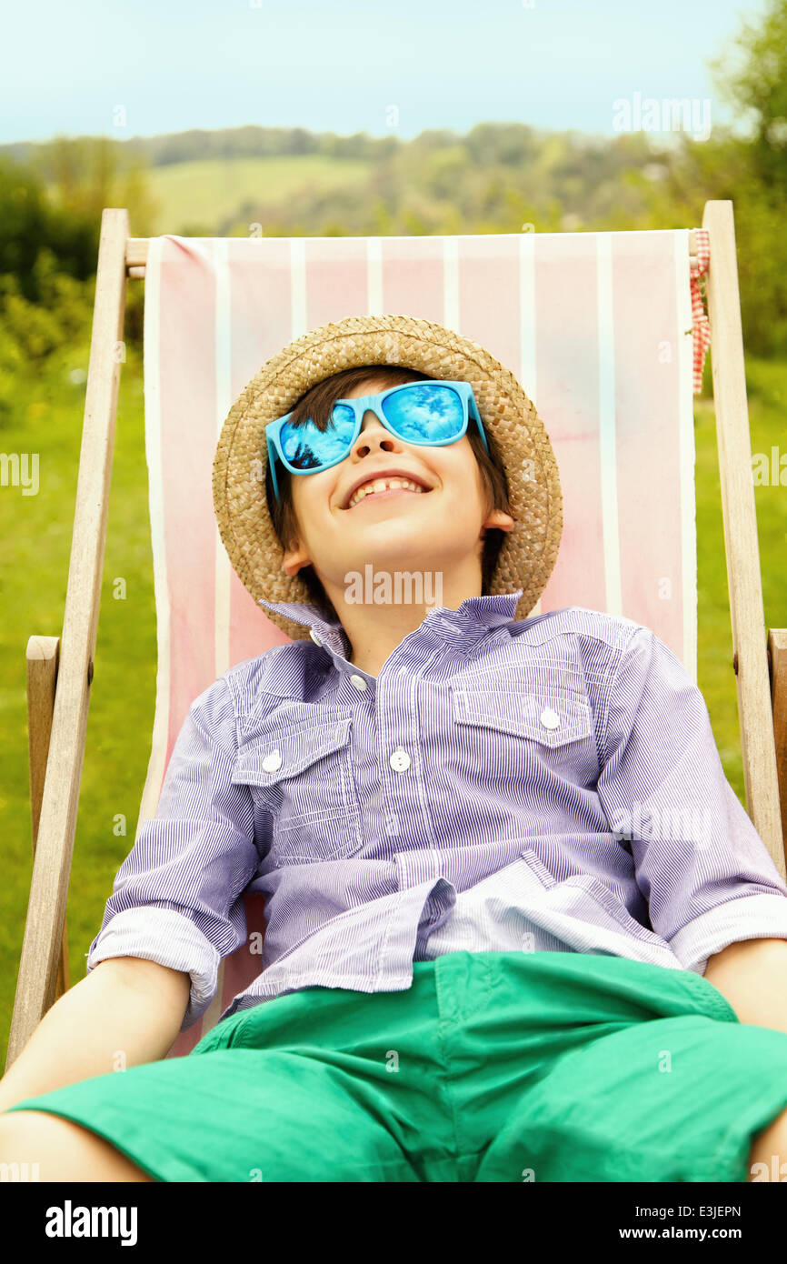 Sonriente niño usando sombrero de paja y gafas de sol sentado en la silla de cubierta Foto de stock