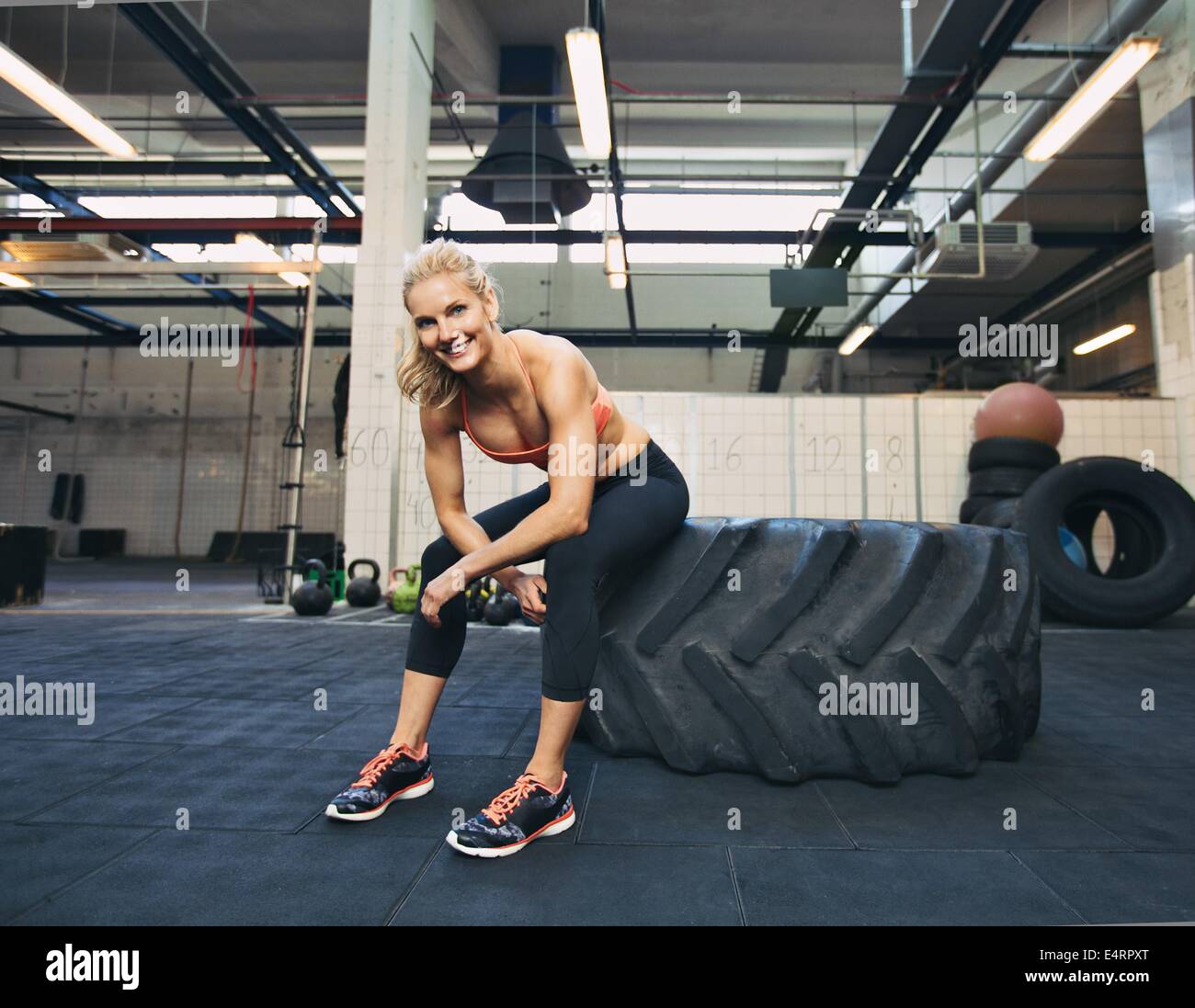 Mujer sentada en el neumático y sonriendo a la cámara en el gimnasio. Crossfit atleta femenina teniendo descanso después del trabajo. Foto de stock