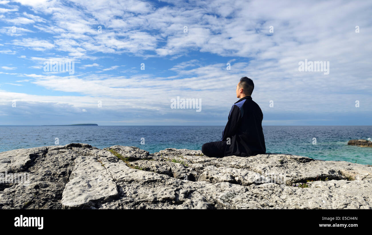 Persona meditando sentado sobre una roca en una orilla de un lago. Península de Bruce, en Ontario, Canadá. Foto de stock