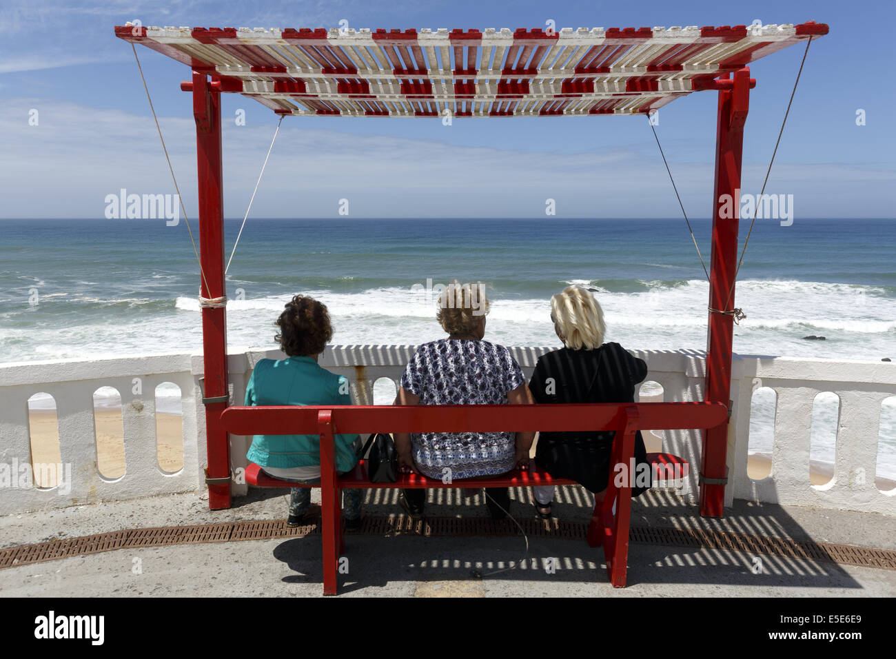 Tres mujeres sentadas en un banco de sombra mirando al mar Silverira, Portugal Foto de stock