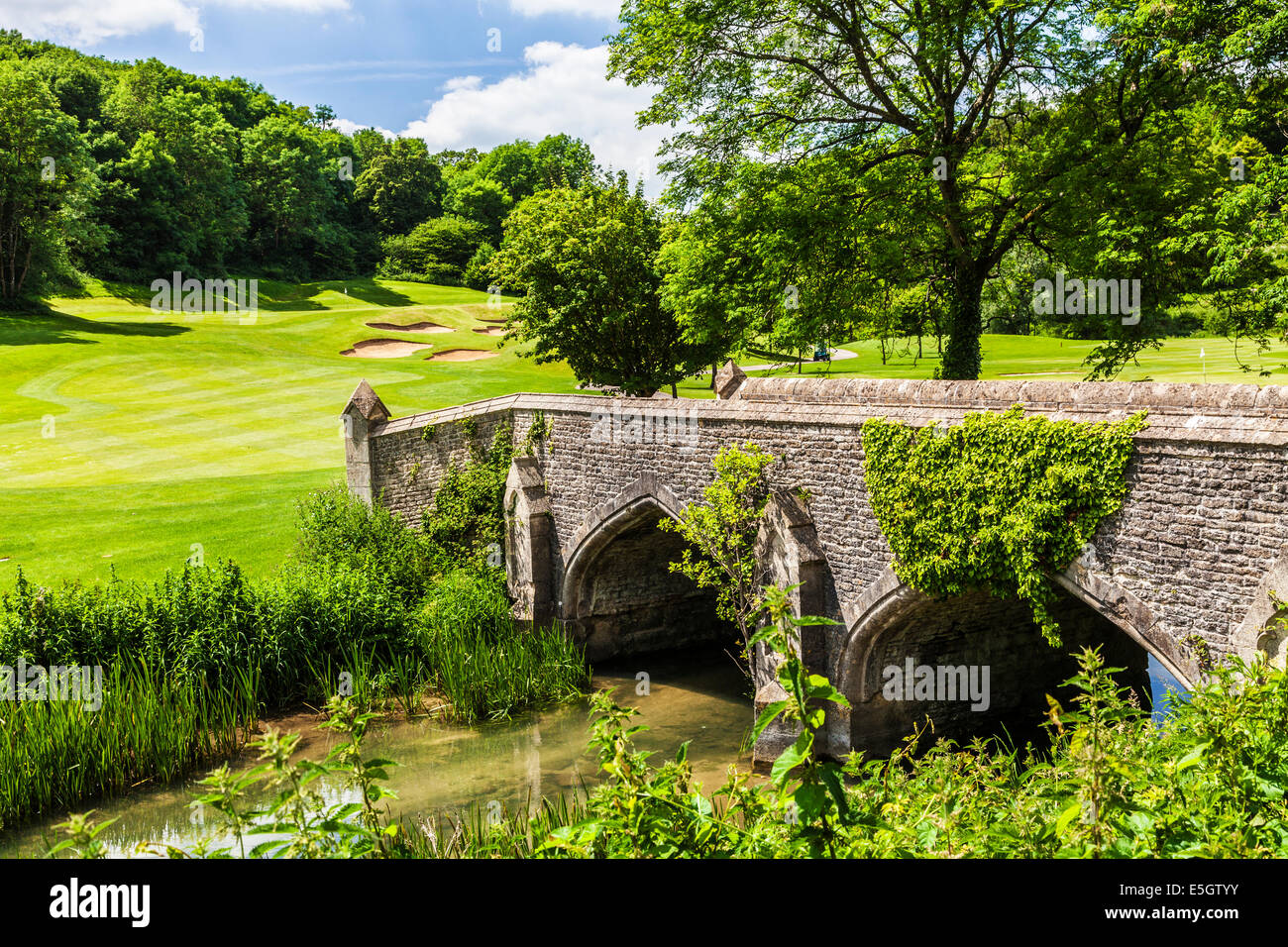 Un puente de piedra sobre el río por arroyo con un campo de golf más allá. Foto de stock